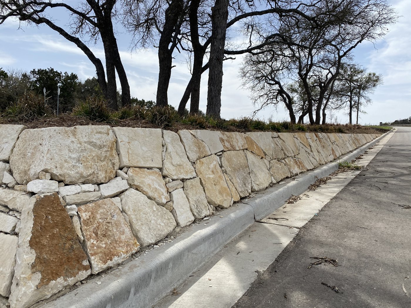 Long random-rubble retaining wall in cream and tan limestone running along a curved roadway under mature oaks