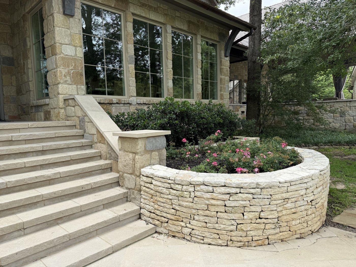 Curved cream limestone planter wall in tight stacked pattern framing a rose garden by a stone-clad residence