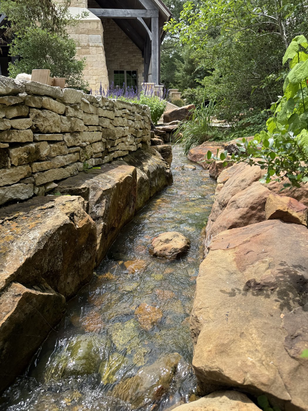 Water feature — narrow stream channel between large rust boulders and a stacked stone wall flowing past a stone-clad home