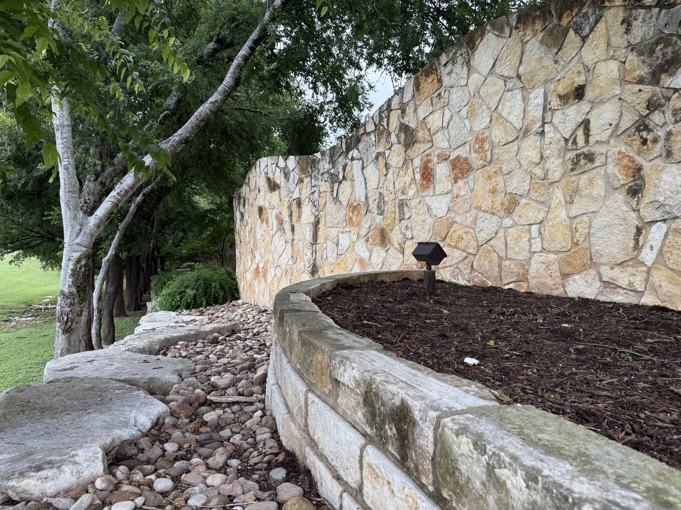 Curving cream and gold random-shape mosaic stone wall with a chiseled cap and natural limestone boulders along the bed below