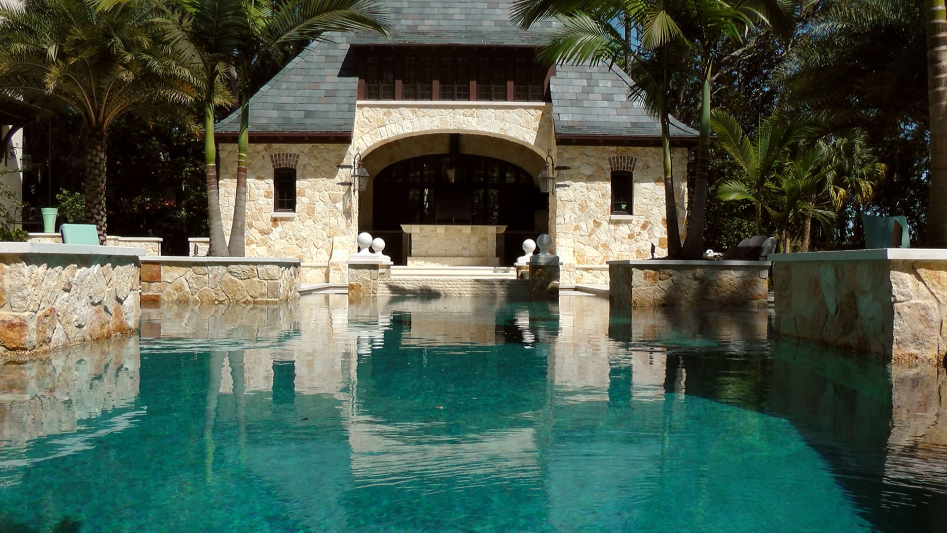 A pool with a large stone house and palm trees.